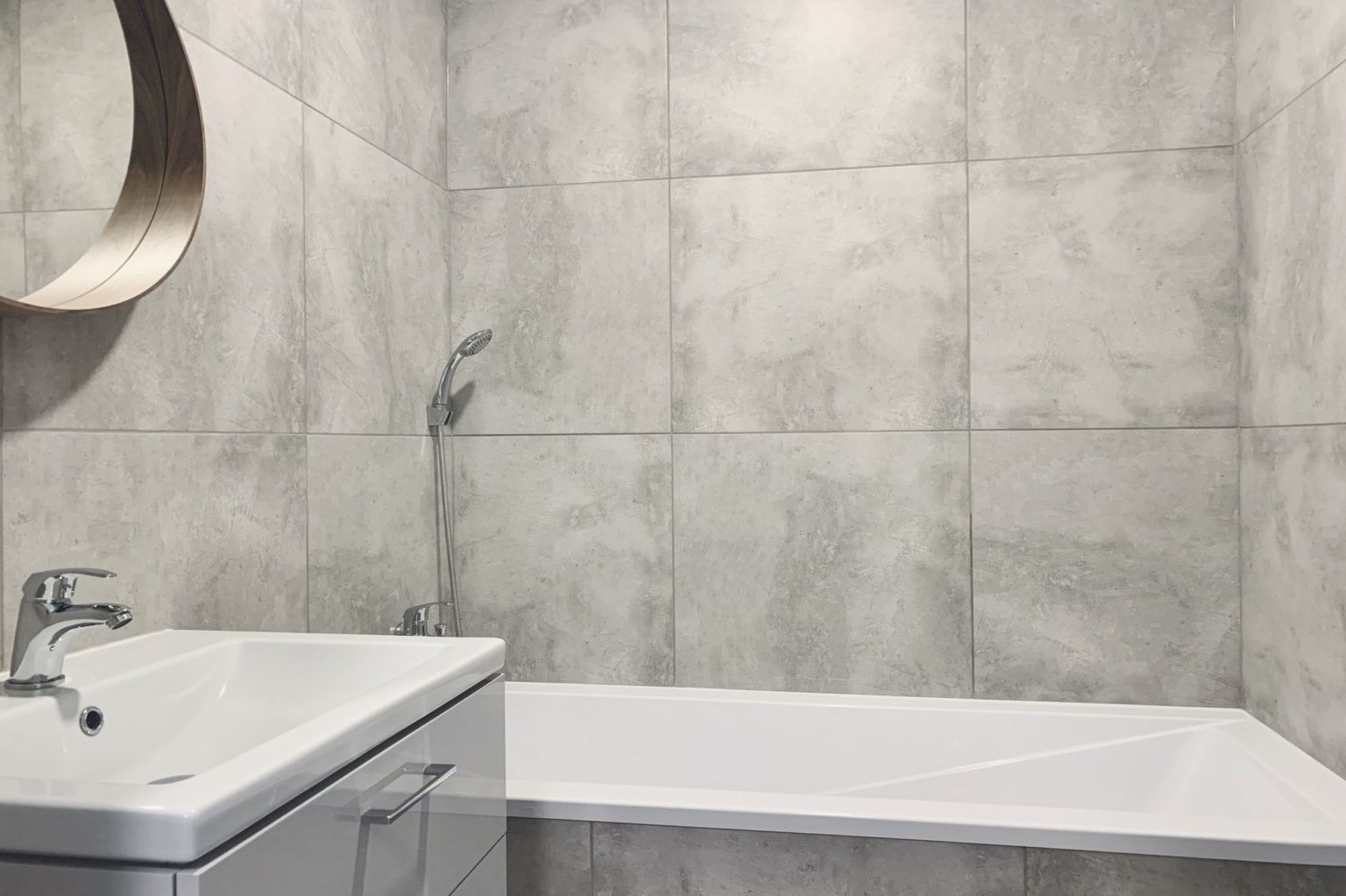 koupelna s šedým obkladem, umyvadlo a vana, kulaté zrcadlo po rekonstrukci